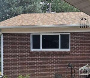 A brick house with a rectangular window, tan shingle roof, and some outdoor items like a wind chime and metal plant stand visible in the foreground. Trees can be seen in the background.