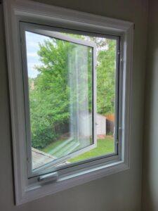 A white-framed window is partially open, showing a view of a green backyard with trees, a wooden fence, and a white shed on a sunny day. The window is set in a light-colored wall.