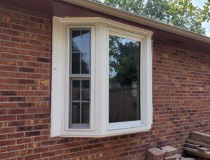 A brick house exterior with a white-framed bay window featuring one vertical pane with grids on the left and a larger, clear pane on the right; stacked bricks lie on the ground nearby.