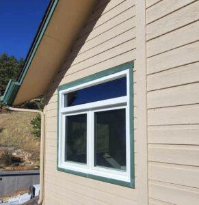 A close-up of a beige house exterior with horizontal siding, showing a white-framed double window topped by a smaller horizontal window. Blue sky and trees are visible in the background.