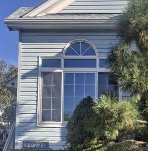 A large window with an arched top on a light blue house exterior is partially shaded by green trees and bushes. A house number sign reading 10526 is visible near the bottom left.