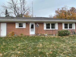 A red brick house with white trim, three windows, and a central glass door. The yard has green grass with scattered leaves, a bush, a wind spinner, and some small plants near the house. Trees with autumn leaves are in the background.