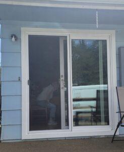 A person sits on a chair inside a house, visible through a glass sliding door. The exterior wall is light blue with white trim, and outdoor patio furniture is seen nearby. Trees and sky are reflected in the glass.