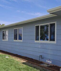 A light blue house with three white-framed windows and a patch of fresh mulch along the foundation. There is a green lawn in the foreground and a small security sign near the right side. The sky is clear and blue.