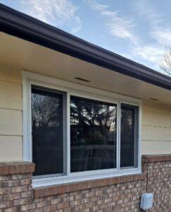 A house window with three glass panels, set in a white frame, is installed in a wall with beige siding and brown brick. Reflections of trees and the sky are visible in the glass.