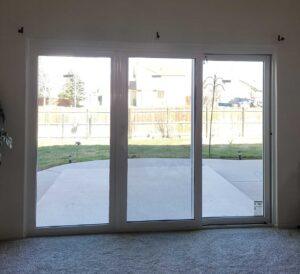 Large glass sliding door in a living room opens to a backyard with a concrete patio, green grass, and a wooden fence. Houses and a few trees are visible beyond the fence under a clear sky.