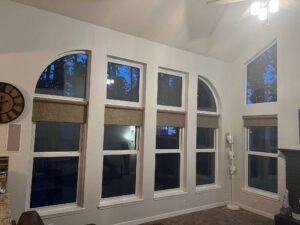 Tall living room windows with white frames in various shapes, including rectangles and arches, with beige shades partially drawn. Blue evening sky and trees are visible outside. A floor lamp is in the corner, and a ceiling fan overhead.