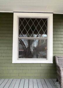 A white-trimmed window with diamond-patterned upper glass is set in a green shingled wall above a gray wooden porch floor. A wicker chair is partially visible on the right. Tree reflections appear in the window.