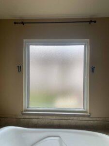 A frosted glass window with a black curtain rod and hooks above a bathtub, set in a tiled bathroom wall with tan-colored paint.