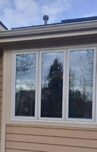 A beige house features three large rectangular windows with white frames, reflecting trees and a cloudy sky. The roof has a small metal vent visible above the windows.