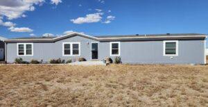 A single-story, gray manufactured home with white trim and multiple windows, set on a dry, brown lawn under a blue sky with scattered clouds.