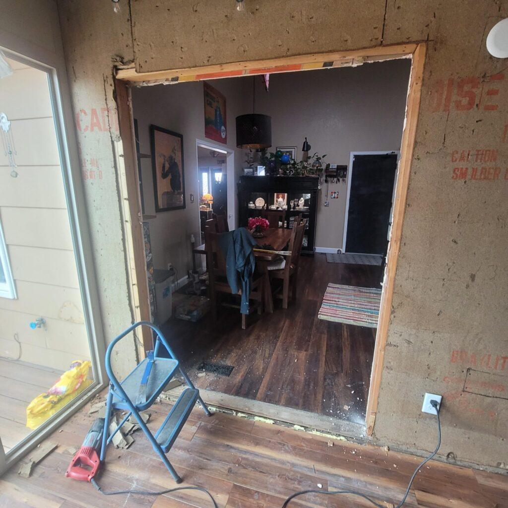 A partial wall renovation reveals an unfinished doorway between two rooms; tools, a step stool, and debris are visible in the foreground, with a dining table and shelves in the background.