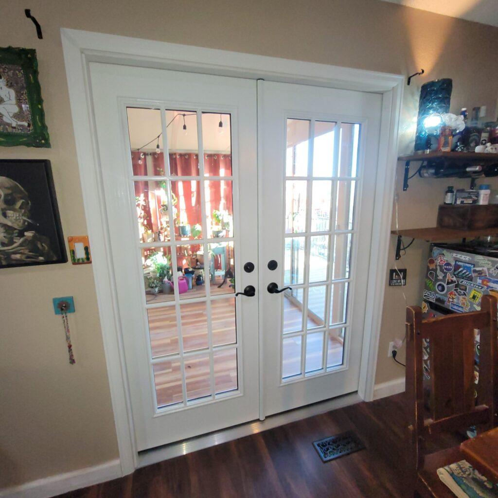 White double French doors with glass panes open to a sunlit room filled with plants. The room has warm wood floors, red curtains, and string lights. The interior space shows art, shelves, and a sticker-covered surface.