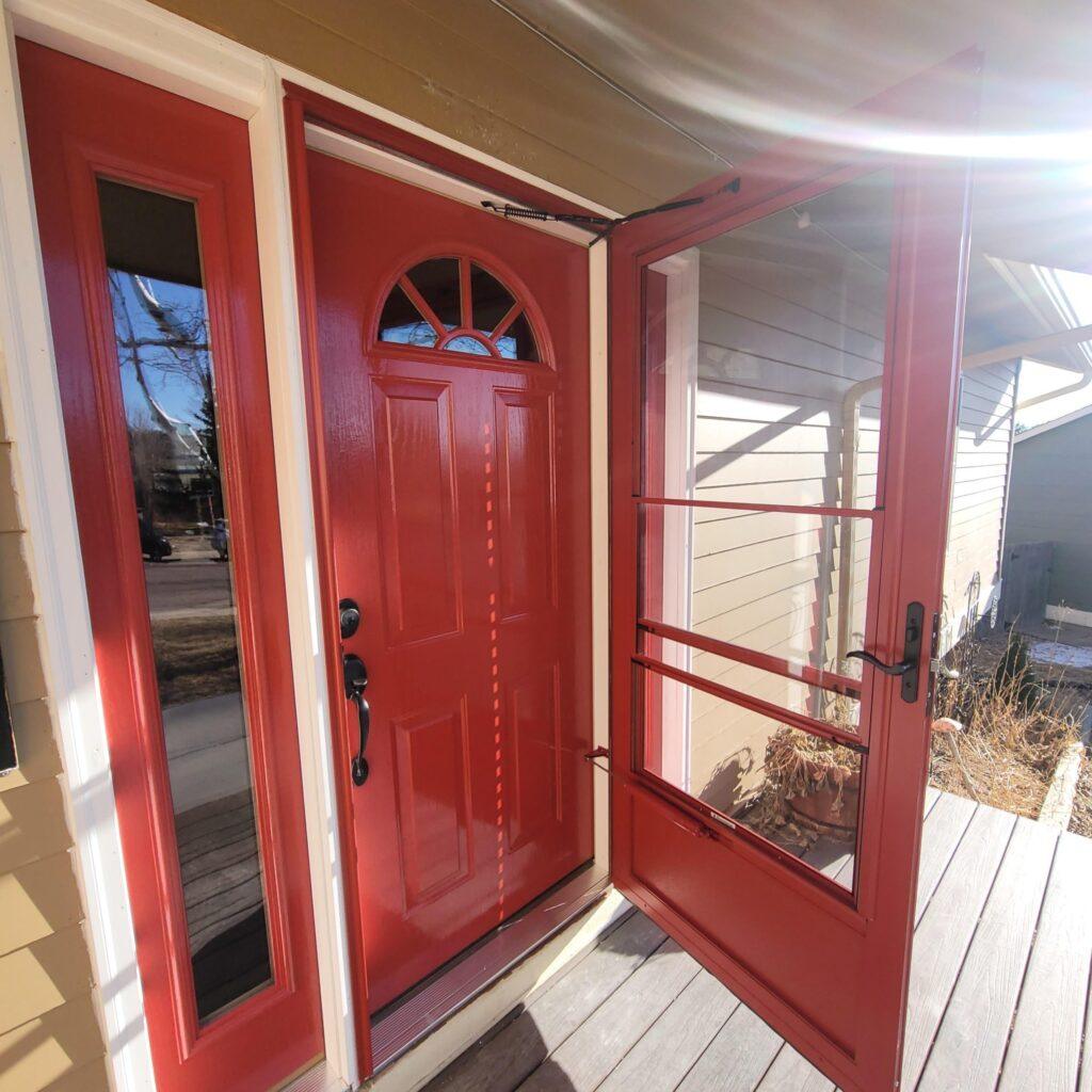 A bright red front door with a matching storm door is open, revealing a porch with wooden boards. Sunlight casts shadows, and a side window reflects outdoor trees and the neighboring house.