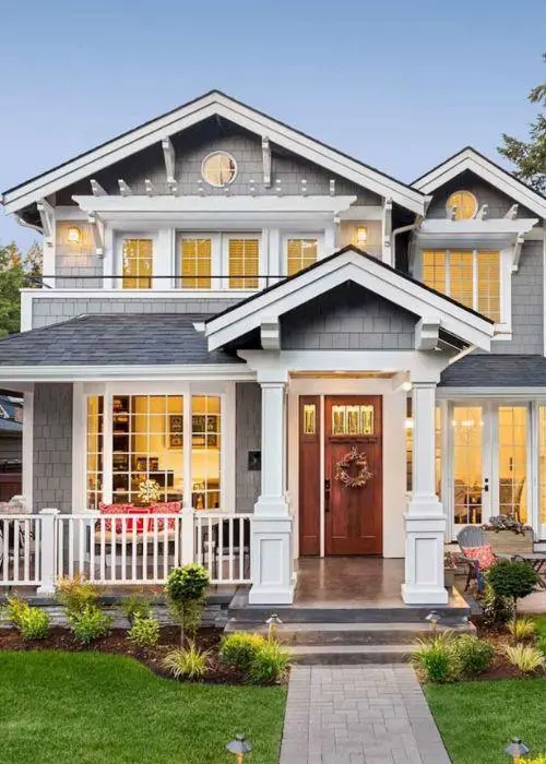 A two-story gray house with white trim, large windows, and a wooden front door decorated with a wreath. The house has a covered porch with white railings and columns, surrounded by green grass and shrubs.