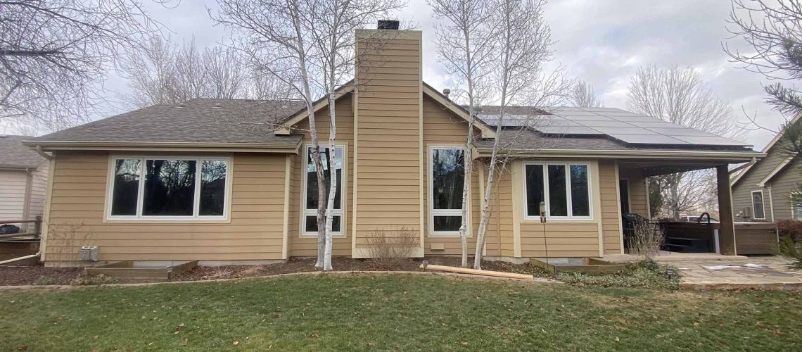 A beige single-story house with large windows, a central chimney, and a solar panel on the roof. Leafless trees surround the house, and there is a green lawn in the foreground under a cloudy sky.