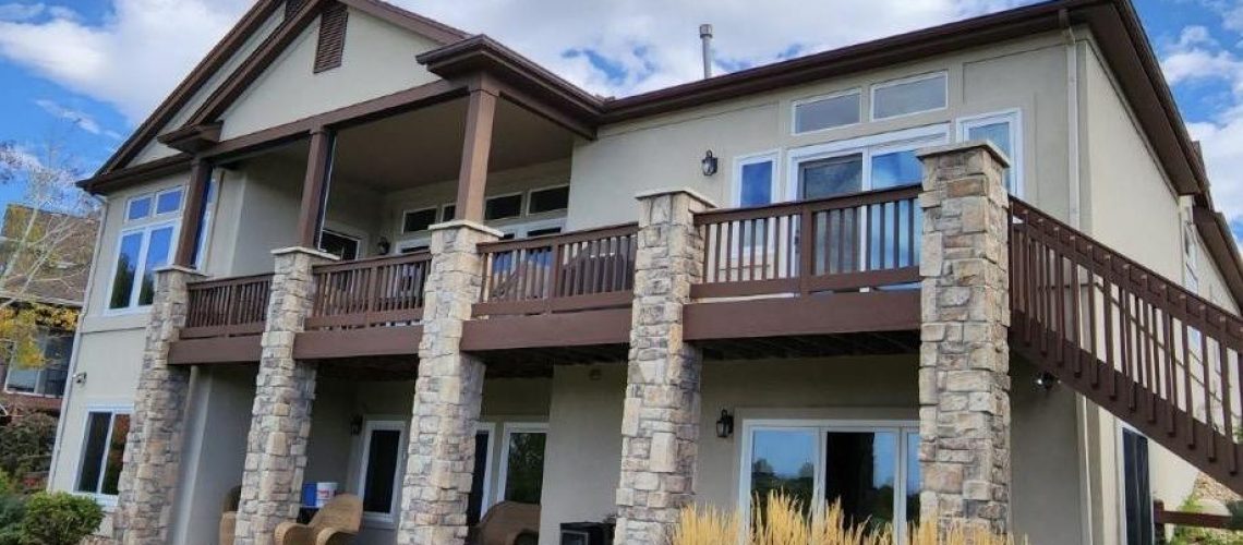 Two-story house with stone columns, beige siding, and a large balcony with wooden railings. Patio furniture sits on the ground level, and tall ornamental grass grows near the building. The sky is partly cloudy.