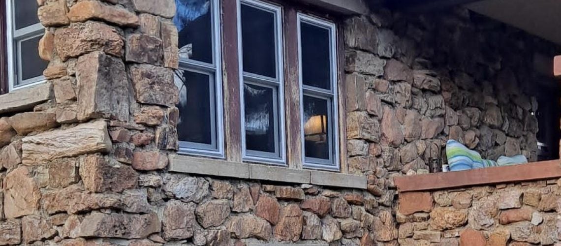 A rustic stone house with multiple windows and a small stone-walled porch, seen under a partly cloudy sky with some pine tree branches overhead.